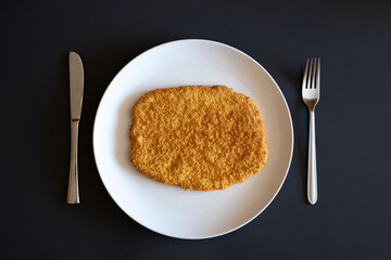 Raw, golden, bread crumb chicken schnitzel on a white plate, knife and fork on the side. Top view, no people