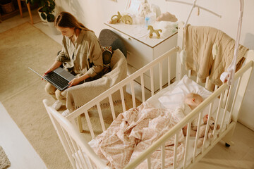 Young woman sitting on chair and working on laptop near baby crib with sleeping baby in cozy room filled with soft light creating relaxing atmosphere