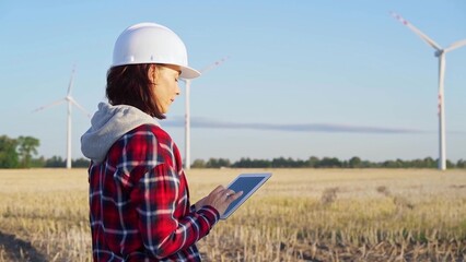 Fototapeta premium Female engineer wearing a white protective helmet is taking notes with a tablet computer on a field with wind turbines, as the sun sets. Clean energy and engineering