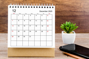 December 2025 desk calendar on wooden table with black smartphone.