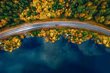 Aerial view of road with red car by blue lake and fall forest with autumn colorful trees.