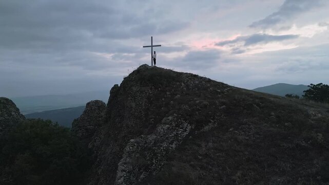 Aerial panning view caucasian woman stand on viewpoint mountain hilltop with giant standing cross on dramatic sunset and cloudy evening in nature. Believer search for purpose hope and pray conceptual