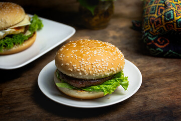 burger with lettuce, cheese, tomato, egg, and beef placed on a white plate with a wooden bottom