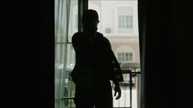 Silhouette of a man in a long-sleeved striped shirt and a cap, standing with a gun raised in front of him, looking menacing, at the entrance of the victim's house.