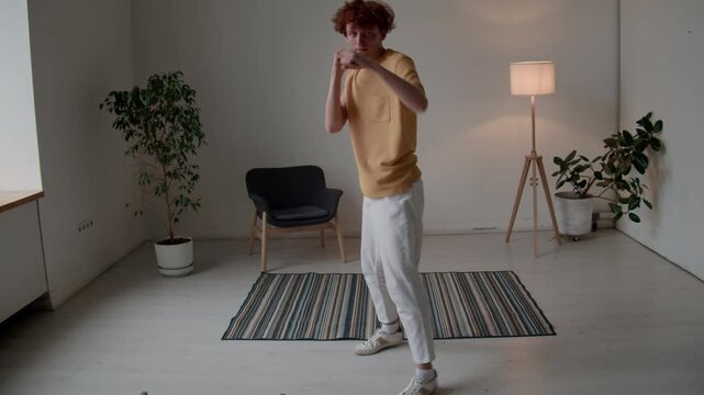POV full shot of young red-haired Caucasian man in casual clothes practicing punches and leg movements while shadowboxing at home alone, looking at camera