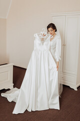 Fototapeta premium A bride is standing in front of a white dress hanging on a hanger. She is wearing a white veil and a necklace. The dress is long and elegant, and the bride is looking at it with a smile on her face