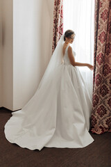 Fototapeta premium A woman in a white wedding dress stands in front of a window. She is wearing a veil and a white dress