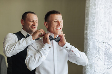 Two men are getting dressed up, one of them is wearing a bow tie. The man in the bow tie is smiling, and the other man is helping him. Scene is lighthearted and fun