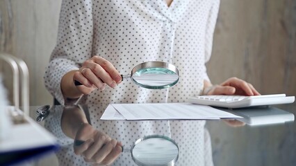 Auditor woman analyzing financial documents with magnifying glass and calculator at her office desk in formal white blouse with polka dots. Business people concept