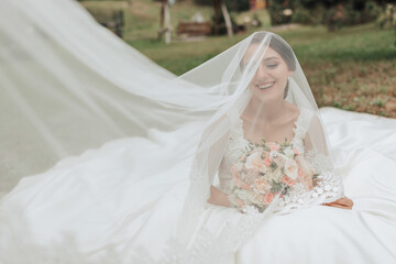 A bride is sitting on a bed with a veil covering her face. She is holding a bouquet of flowers