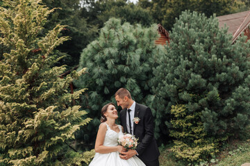 A bride and groom are standing in front of a forest with trees. The bride is holding a bouquet and...