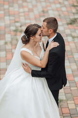 Naklejka premium A bride and groom are kissing on a brick walkway. The bride is wearing a white dress and the groom is wearing a black suit. Scene is romantic and happy
