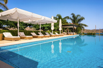 Swimming pool area with rattan sunbeds and large umbrellas, creating comfortable and stylish lounging space. Poolside surrounded by lush greenery, providing natural and serene backdrop.  