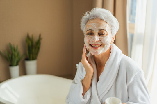 Mature adult old woman applying nourishing anti age face cream doing daily beauty routine. Senior woman with white mask or creme on skin gets facial treatment Skin care aging beauty skincare cosmetics