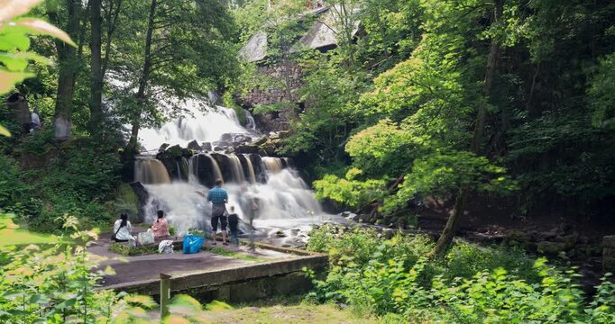 Rottle Waterfalls, Sweden. A picturesque waterfall surrounded by a dense forest. Sunlight peeks through the trees, illuminating the cascading water. Timelapse of visiting people during long exposure.
