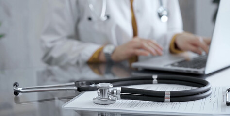 Stethoscope and clipboard with medication history records are on a glass desk behind a physician using a laptop computer. Medicine concept