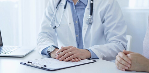 A doctor and a patient. The physician, wearing a white medical coat over a blue shirt, is filling out a medical record form during a consultation in the clinic. Medical service
