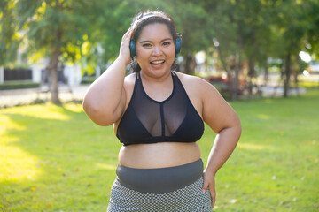 Portrait sportswoman listening music through headphones. beautiful young chubby overweight woman in sportswear doing exercise outdoors at park. fitness sport club, body and health care. Asian female