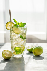 Mojito cocktail in a tall glass with lime, mint and ice on a table with morning light and shadow.
