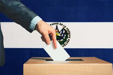 A hand holds a ballot in a ballot box against the background of the El Salvador flag. People cast...