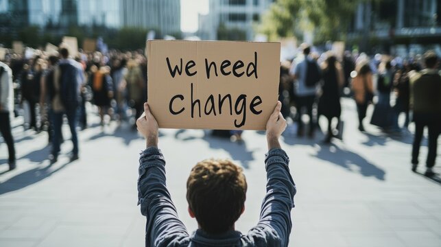 A person holding a sign with the text "we need change" at a peaceful protest, a crowd of people in the background in a city square.