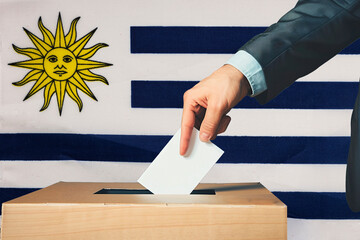 A hand holds a ballot in a ballot box against the background of the Uruguay flag. People cast...