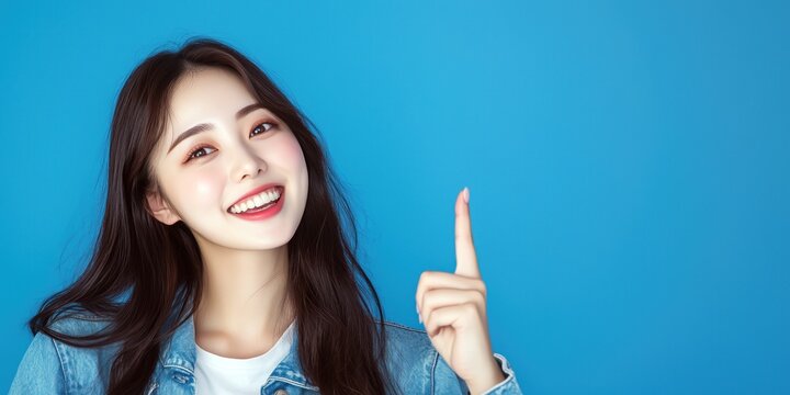 Une jeune femme asiatique brune aux cheveux longs, souriante et pointant son doigt sur un fond bleu cr&eacute;ant un visuel attrayant pour la publicit&eacute;.