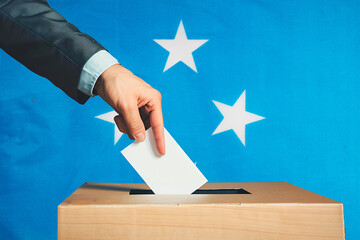 A hand holds a ballot in a ballot box against the background of the Micronesia flag. People cast...
