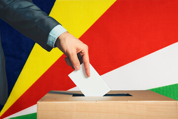 A hand holds a ballot in a ballot box against the background of the Seychelles flag. People cast...