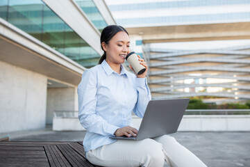 A young Asian woman sits comfortably on a wooden bench, sipping coffee from a cup while focused on...
