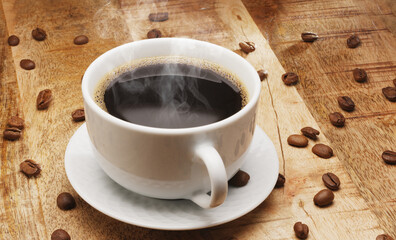 White cup of hot steaming coffee on a wooden table and roasted coffee beans near it.