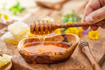 Honey dripping from wooden spoon into wooden bowl with honey, spring flowers are on wooden table...