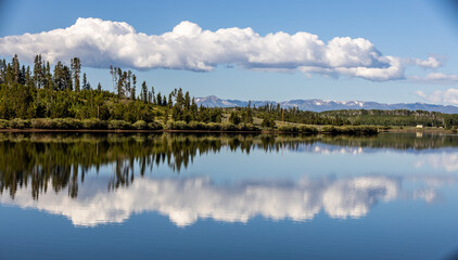 Obraz premium reflections of pines and clouds across a clear body of water with mountains in the distance