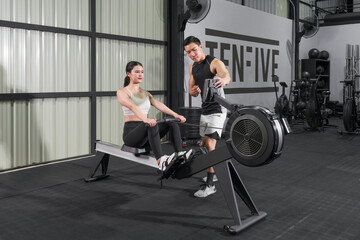 An athletic woman in sportswear is engaged in an intense workout session on a rowing machine with a trainer inside a well-equipped gym. © chadchai