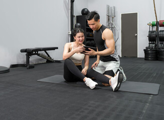 Obraz premium Young asian female sitting on the rubber floor in the gym, check the exercise schedule through tablet computer with male instructor.