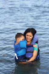 Obraz premium A asian mother holds her son, they are ware a life jacket while playing in the sea.