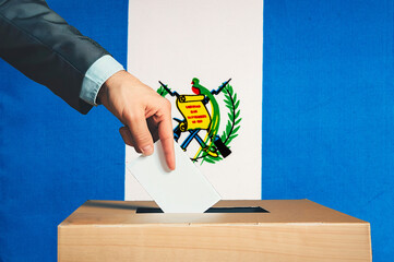 A hand holds a ballot in a ballot box against the background of the Guatemalan flag. People cast...