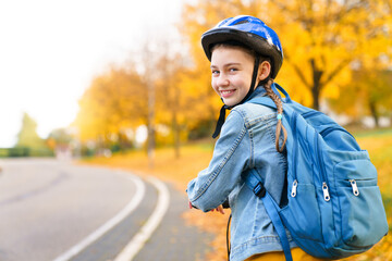 School child in protective helmet on safety road, cycle lane. Happy kid riding scooter on autumn city street. Girl with blue backpack smiling. Back to school. Sustainable transportation and lifestyle