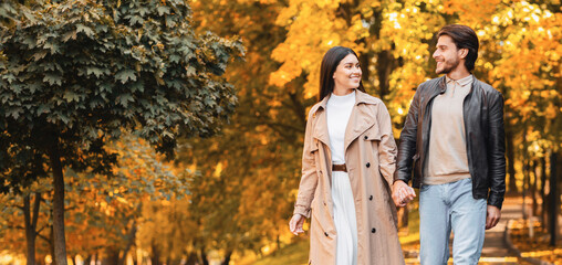 Positive man and woman holding hands, talking and walking by golden park, empty space