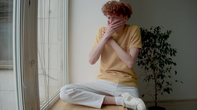 Full shot of young Caucasian guy in t-shirt and pants sitting on wooden window sill and looking at disgusting situation outdoors, feeling nauseous