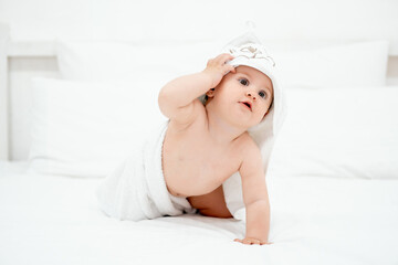 a baby in a towel on a white bed at home, a smiling child after bathing in a bright bedroom, a space for text