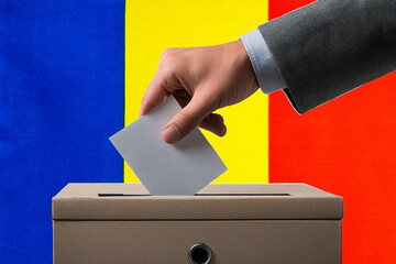 A hand holds a ballot in a ballot box against the background of the Romanian flag. People cast...