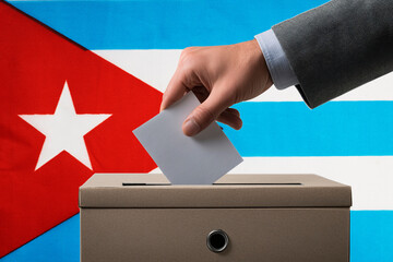 A hand holds a ballot in a ballot box against the background of the Cuban flag. People cast ballots...