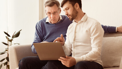 Mature man teaching old dad to use laptop, computer programs and browse internet sitting on sofa...