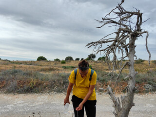 Thirsty woman in a yellow shirt hiking and reaching by the dry tree in dried land of nature park Kamenjak in Premantura, Croatia