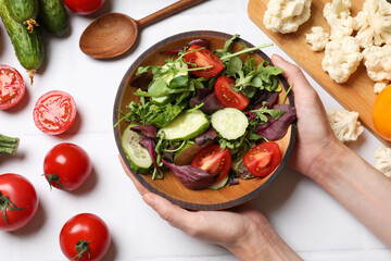 Healthy vegetarian food. Woman holding bowl of salad at white tiled table with vegetables, top view