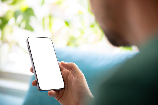 Person holding a smartphone with a blank screen, relaxing on a sofa in a bright, green environment. Perfect image for presenting mobile apps, user interfaces, or design templates.