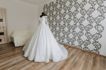 A long white gown is displayed in a room with a wall of floral wallpaper. The gown is draped over a...