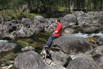 A man waits for his mongrel dog to quench his thirst in a small mountain river in Norway.
