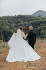 A bride and groom are walking together in a field. The bride is wearing a white dress and the groom...